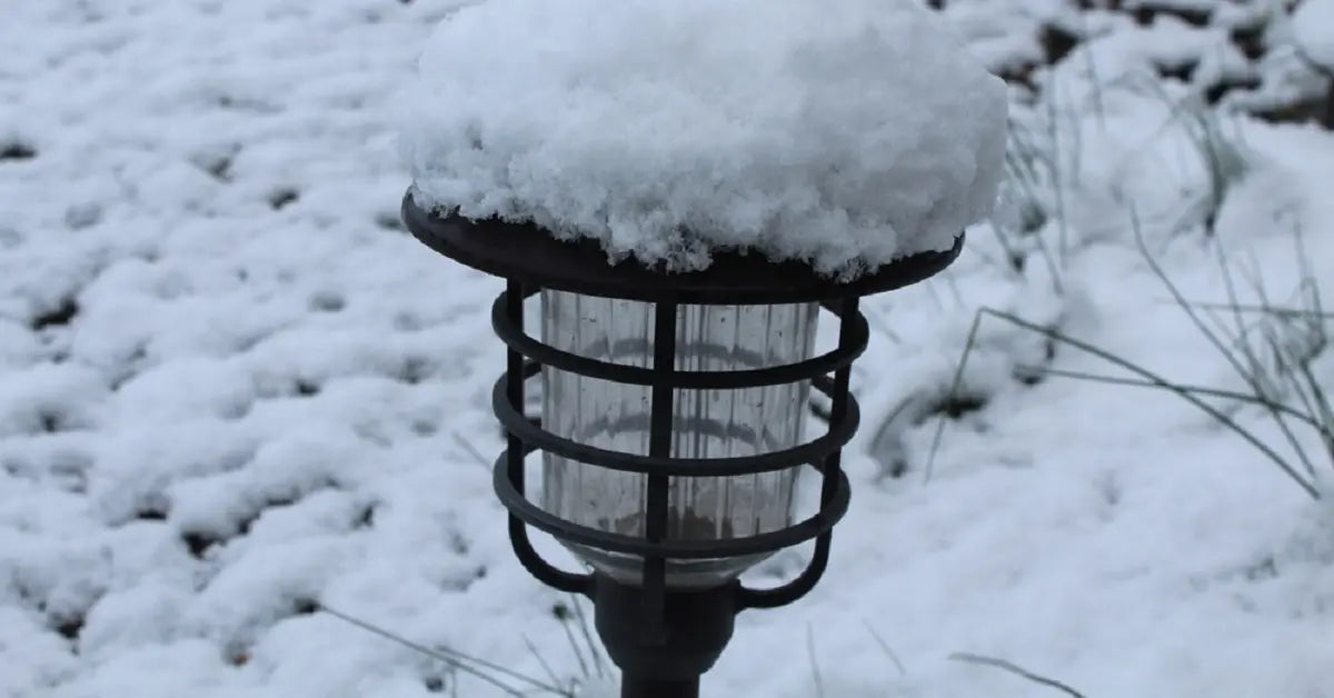 A black solar garden light with a thick layer of fresh snow on top, standing in a snow-covered garden bed with sparse grass peeking through.