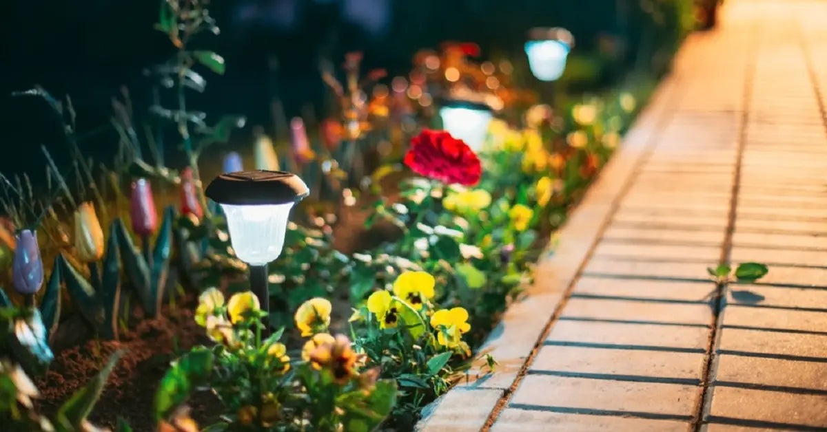 Solar-powered pathway lights illuminating a flower bed along a stone walkway at dusk, highlighting garden plants and blooms.