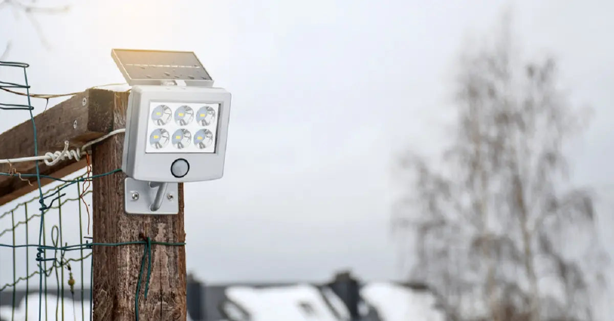 A small solar-powered security light mounted on a fence post in a snowy, overcast winter scene, showing that solar lights are designed to work in winter conditions.