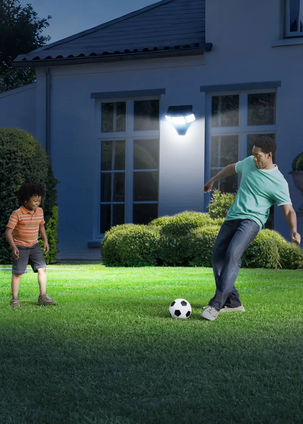 Vertical photo intelamp solar motion‑sensor wall light illuminating home backyard lawn at night, father and child playing football, solar security lighting for yard