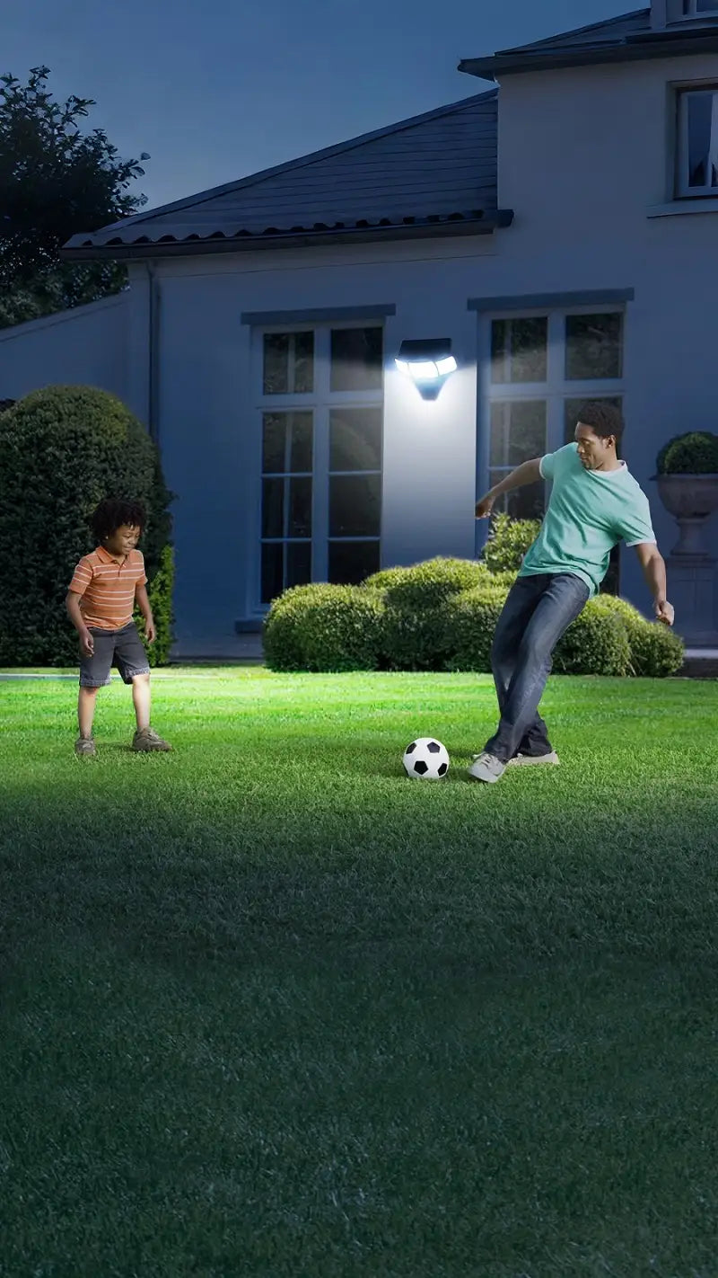 Vertical photo intelamp solar motion‑sensor wall light illuminating home backyard lawn at night, father and child playing football, solar security lighting for yard